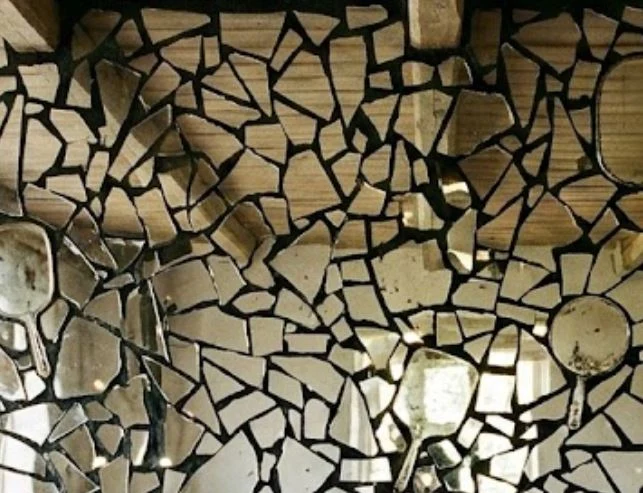 A wall covered in a mosaic of broken mirror fragments set in dark mortar, with old hand mirrors embedded among the pieces, reflecting a room filled with light.